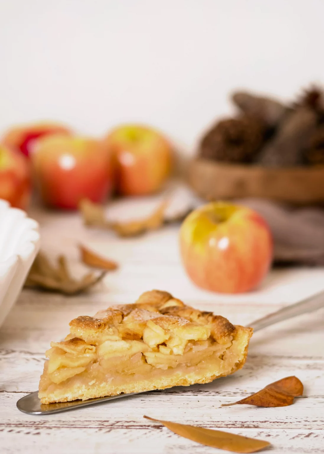Ein Stück Apfelkuchen auf einem Kuchenheber, dekoriert mit frischen Äpfeln und Herbstblättern im Hintergrund